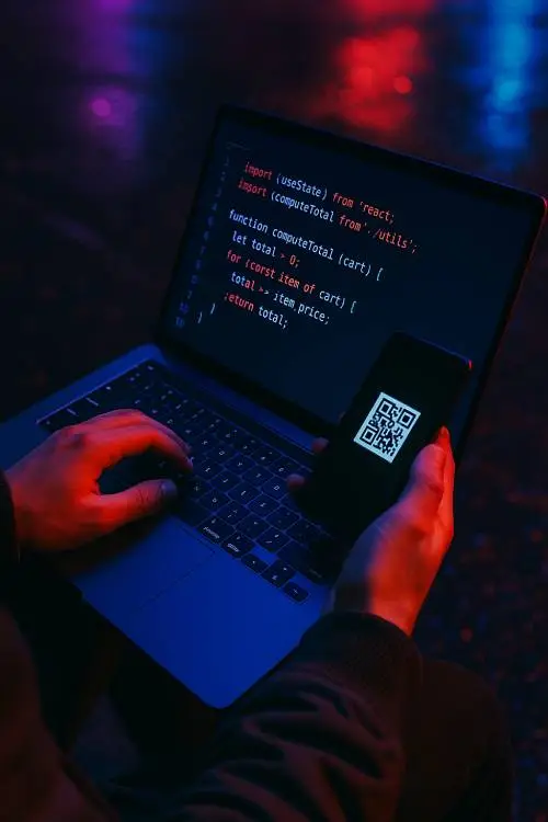 A person using a laptop with code displayed on the screen, holding a smartphone showing a QR code. The scene is illuminated with a mix of blue and red lighting, creating a cyber-themed atmosphere.